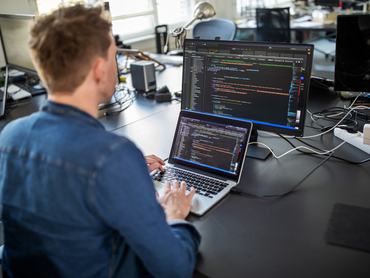 Businessman working on laptop and looking at desktop computer monitor. Male professional sitting at his office desk and working on new software program.