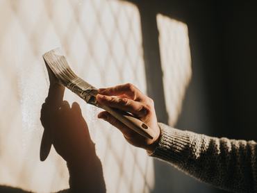 Sunlight shines through a window, creating a sun beam on an interior wall. A man holds a paintbrush and paints the wall a neutral colour. Simple and conceptual. The shadow and wall provides a space for copy.