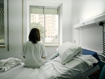 sick woman alone sitting on the bed of a hospital room looking out the window seen from behind
