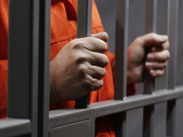 Hands close up of elderly prisoner in orange uniform holding metal bars, standing in prison cell. Criminal serves imprisonment term for crime. Inmate in jail or correctional facility. Justice system.