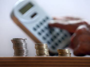 ILLUSTRATION - 15 May 2024, Bavaria, Kaufbeuren: ILLUSTRATION A hand holds a calculator behind three stacks of euro coins. Photo: Karl-Josef Hildenbrand/dpa (Photo by Karl-Josef Hildenbrand/picture alliance via Getty Images)