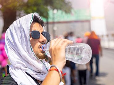 Close-up of a young man covering his head with a cloth and wearing black sunglasses on an extremely hot afternoon while drinking water from a plastic bottle.