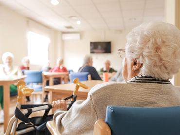 Back view of senior woman with short white hair sitting on chair with foldable walker with friends in blurred background at nursing home