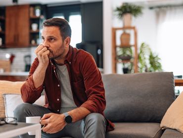 Worried man sitting on the sofa at home, biting fingernails while holding a cup of coffee, deeply lost in thought and grappling with feelings of anxiety and uncertainty