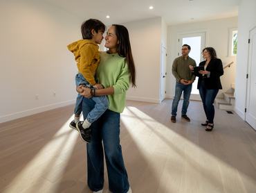 A a mom and son smiling at each other in a new modern home while a man speaks to a real estate agent in the background