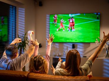 Candid shot of mid-aged parents and young daughter sitting on brown sofa, arms raised in celebration while watching soccer match on large-screen television.
