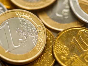 A detailed, top-down view of a pile of Euro coins. The focus is sharp on a two-Euro coin in the center, surrounded by other gold-colored coins. This image can represent the European economy, finance, currency, and wealth.