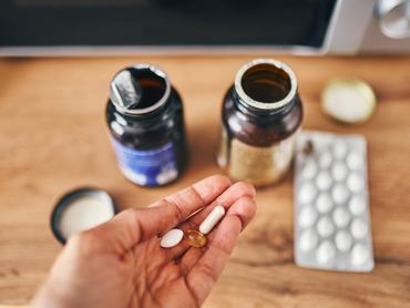 A close-up of a hand holding assorted pills and capsules, with medicine bottles and a blister-pack on a wooden desk, conveying health, care, and self-management in a home or clinical setting.