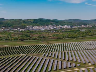 An expansive solar farm operates in the foreground with a large industrial plant and lush green hills under a bright blue sky.