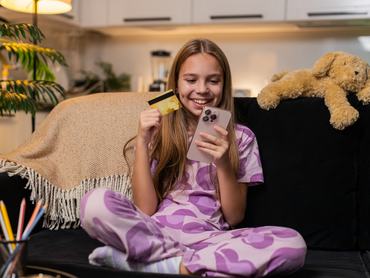 Teenager girl uses smartphone with credit card at night home picks book or toy online smiles happy. Caucasian child kid on sofa at evening adds to cart checks pay form types info taps confirm order