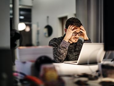 Young casual businessman wearing glasses is sitting in front of his notebook holding his head pondering over his work. Office equipment and another computer is in front of him.