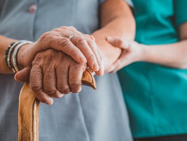 Photo of Unrecognizable Senior Woman's Hands On Walking Stick With Care Worker In Background. Young woman holding hand of old woman with walking stick. Female nurse supporting senior woman in with walking stick at home during the day.