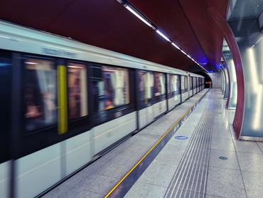 A sleek subway train captured in motion blur speeds through a contemporary underground station. The futuristic platform features a distinctive burgundy ceiling with recessed lighting, clean granite flooring with tactile guidance strips, and a minimalist design that exemplifies modern urban transportation infrastructure.